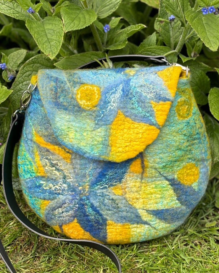 Nuno Felted Cornflower Blue Handbag Made by Liz Riley Nuno Felted Handbag is a one of a kind art piece and is wet felted using blended Merino wool and a colourful selection of hand dyed silks. It is elegant, lightweight and a great medium sized handbag with a useful inside pocket.  This method of felting uses soap, warm water and agitation to tangle fibres together to create felt. This bag is made around a resist (pattern) in one piece so completely seamless, small nickel D rings are felted into the bag to attach a lovely soft black leather strap.