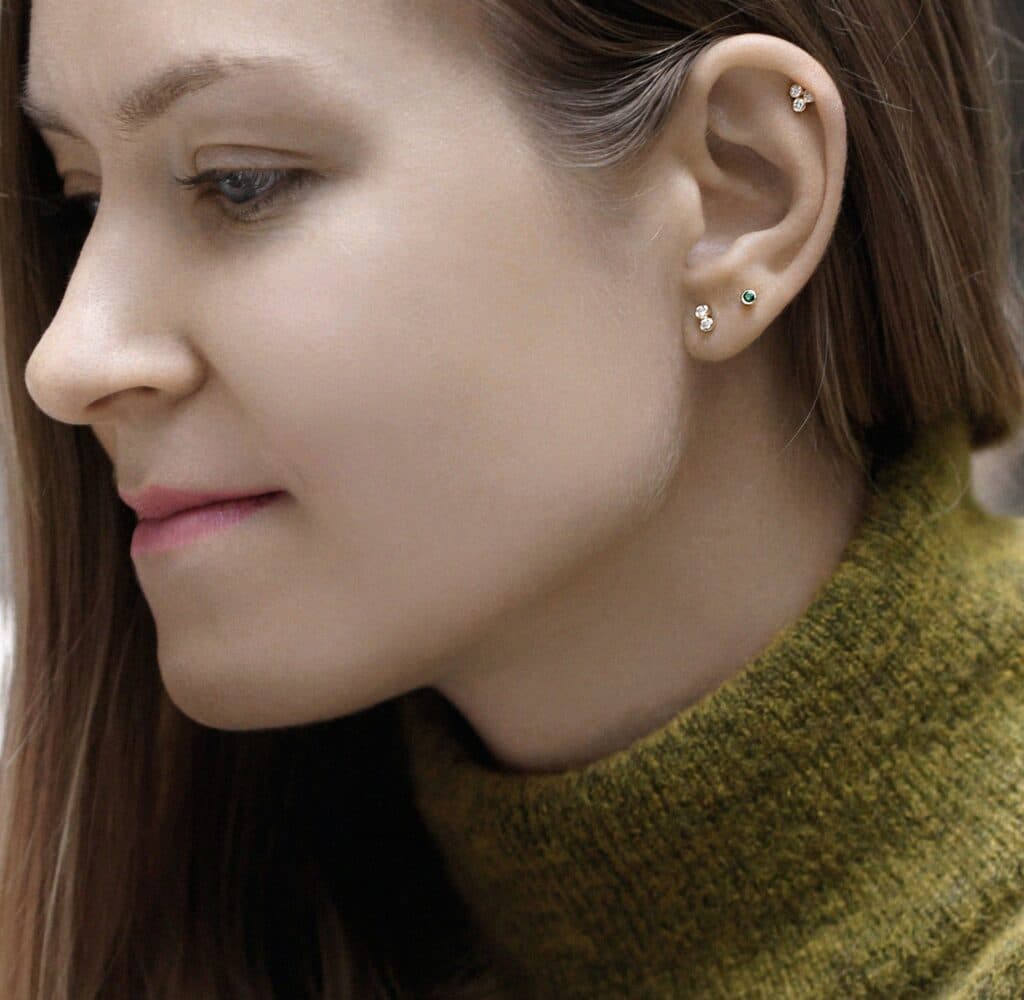 Curated ear styling with emerald and diamond piercing studs in 18k gold. Lena Cohen Jewellery London handcrafted fine piercing jewellery, quiet luxury everyday look.